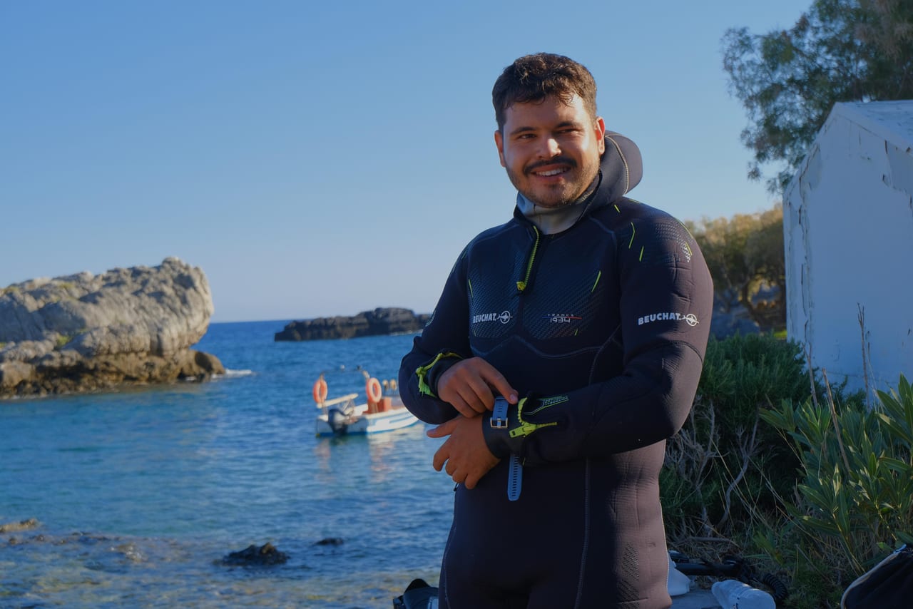 Giannis in a wetsuit at the shore, with the dive boat behind him